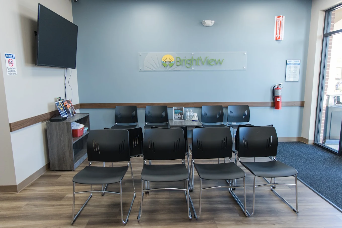 BrightView Health lobby with rows of black chairs, branded wall sign, television, informational materials, and entrance doors