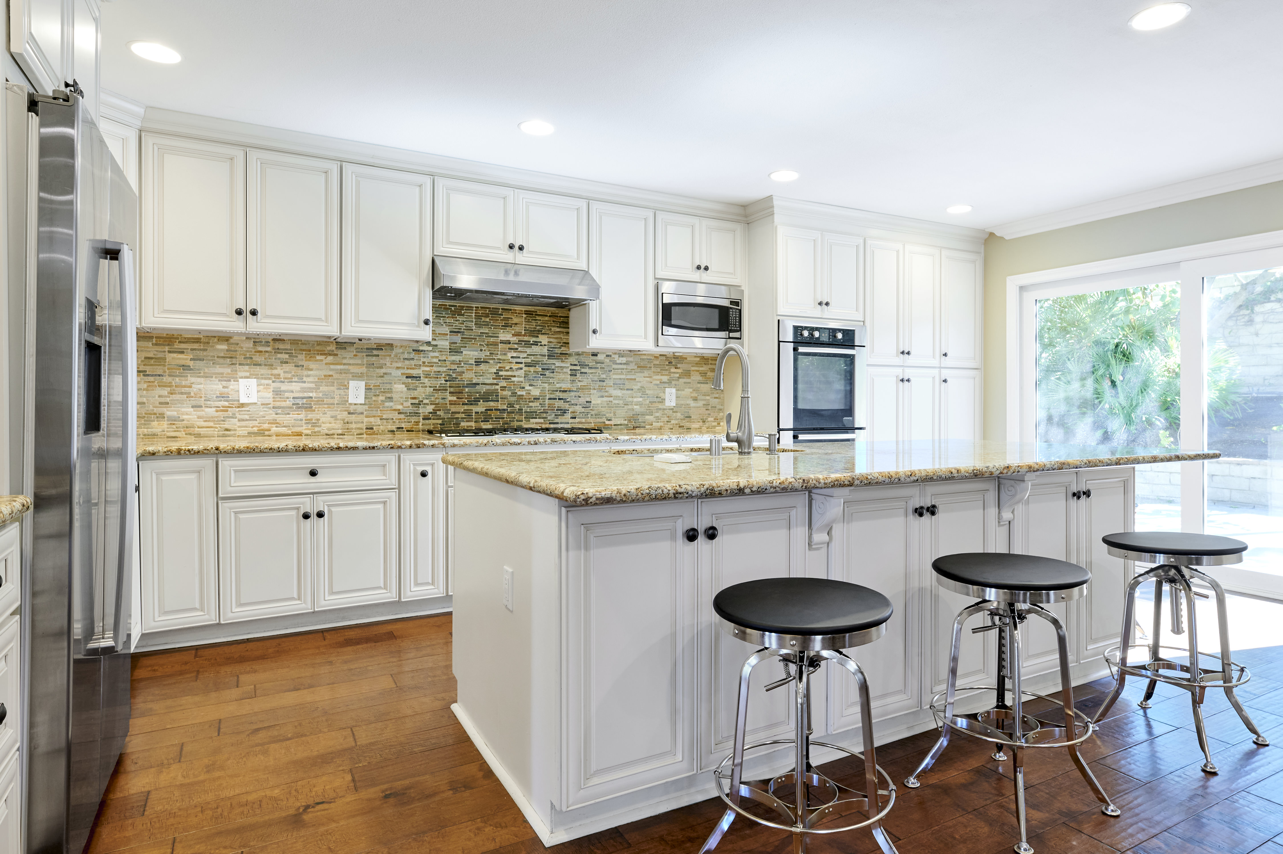 A bright, modern kitchen featuring white cabinets, granite countertops, a stainless steel fridge, and an island with barstools, offering ample space.
