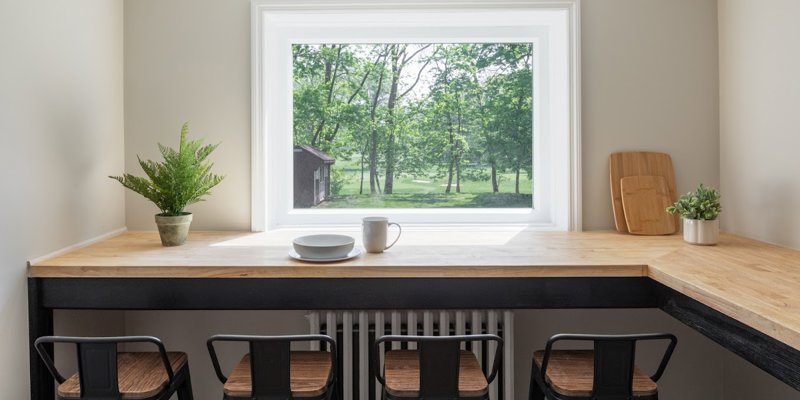 A cozy breakfast nook with a wooden countertop, modern stools, and a large window offering a peaceful view of trees and greenery.