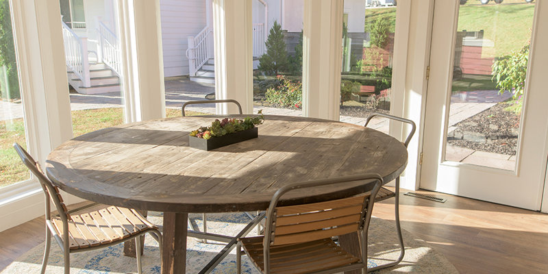 A bright, inviting sunroom features a rustic wooden round table with four chairs, surrounded by large windows that offer scenic outdoor views.