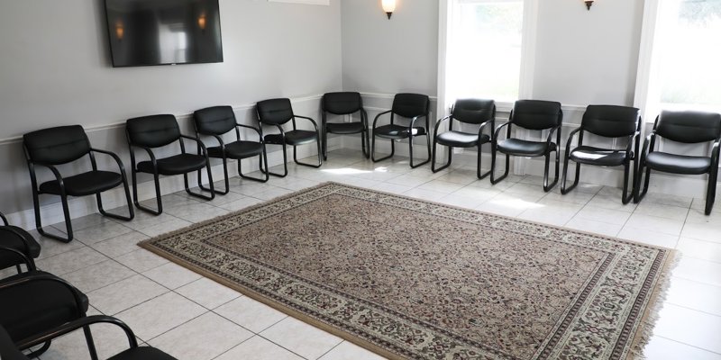  A clean, spacious meeting room with black chairs arranged in a circle, a large area rug, and natural light streaming through the windows.