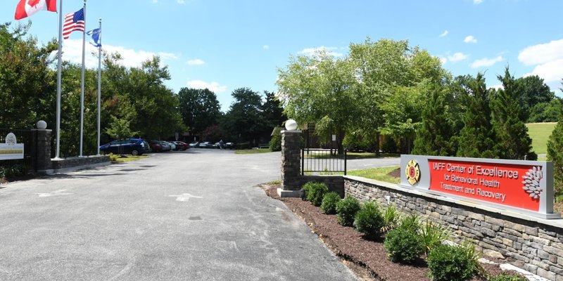  A well-maintained entrance to the IAFF Center of Excellence for Behavioral Health Treatment and Recovery, with flags and landscaped greenery along a wide driveway.