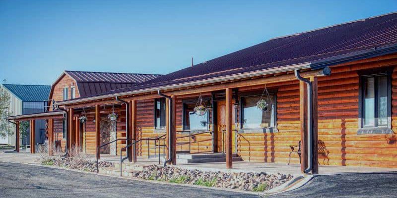  A rustic log cabin-style building with a charming front porch, hanging planters, and a warm, inviting atmosphere under a bright blue sky.