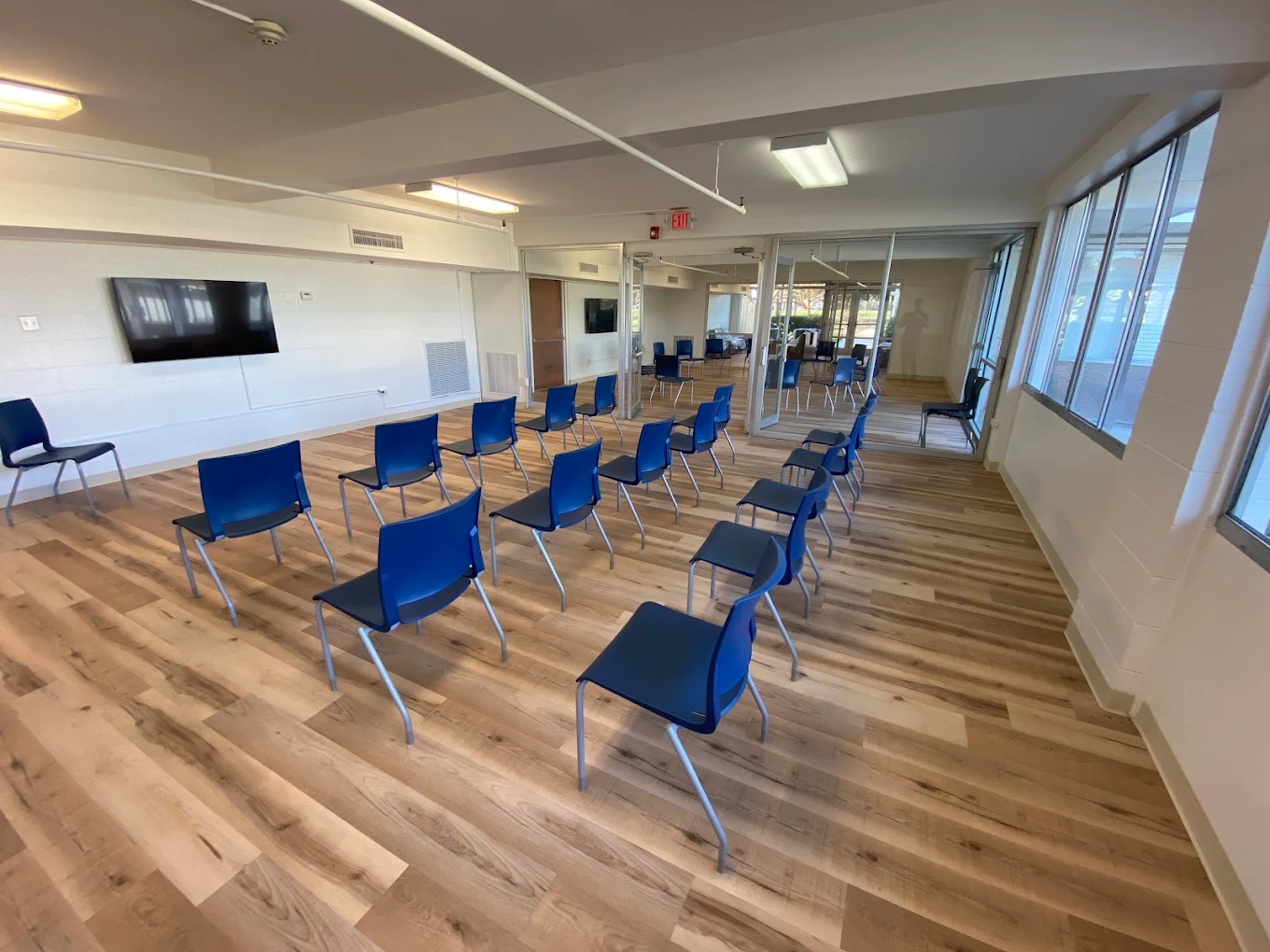 Bright meeting room with rows of blue chairs, wood flooring, wall-mounted television, and large windows