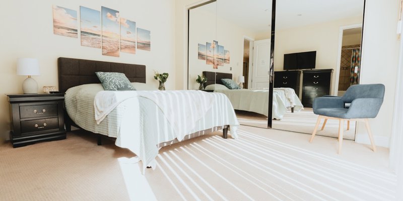 A bright, inviting bedroom with a comfortable bed, stylish decor, and ample natural light reflecting off mirrored closet doors. Perfect for relaxation.