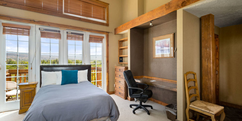 A cozy bedroom with warm wooden accents, large windows letting in natural light, and a serene view. Includes a workspace nook.