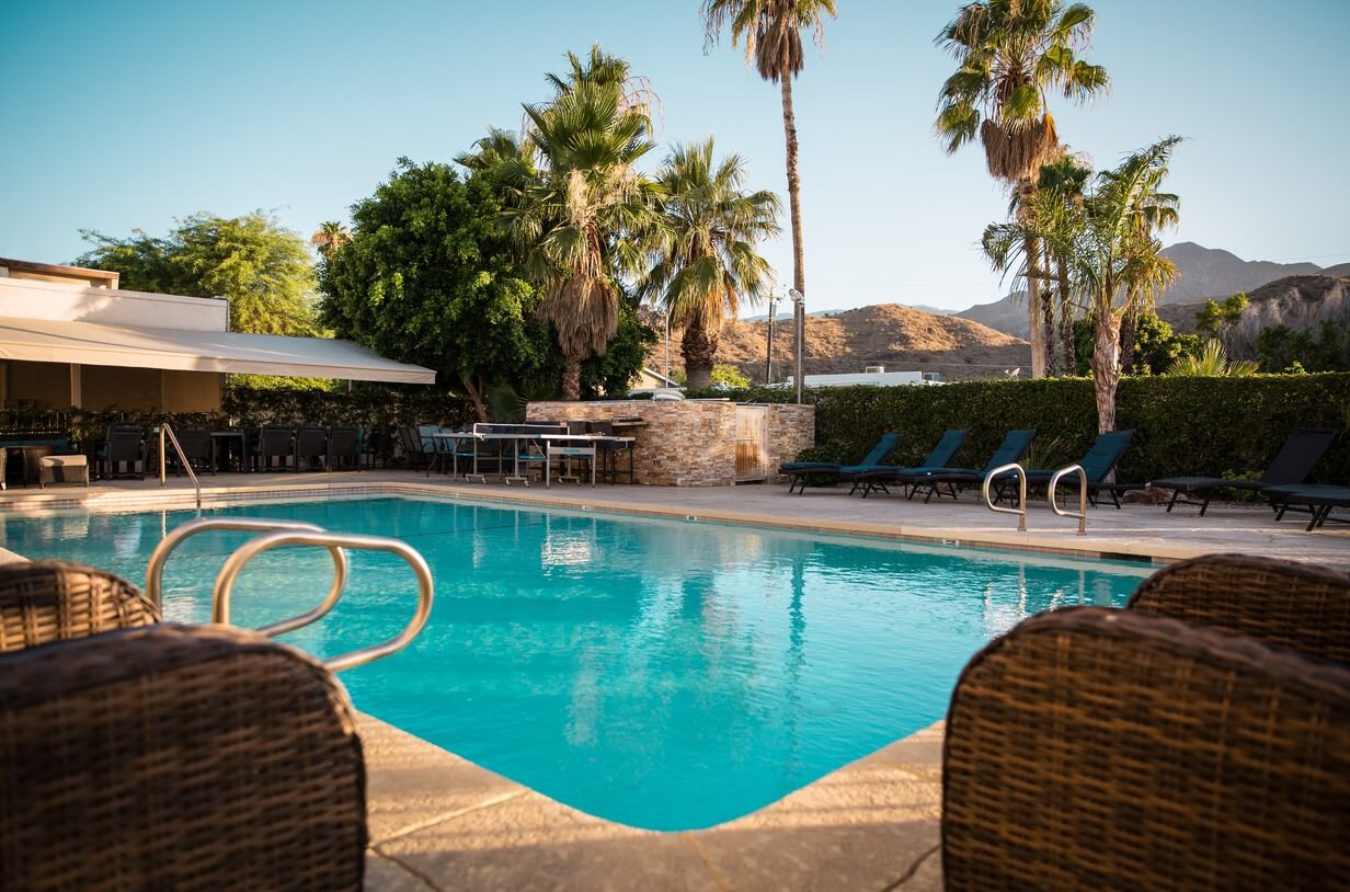 Outdoor pool surrounded by lounge chairs, palm trees, and mountains in the background.