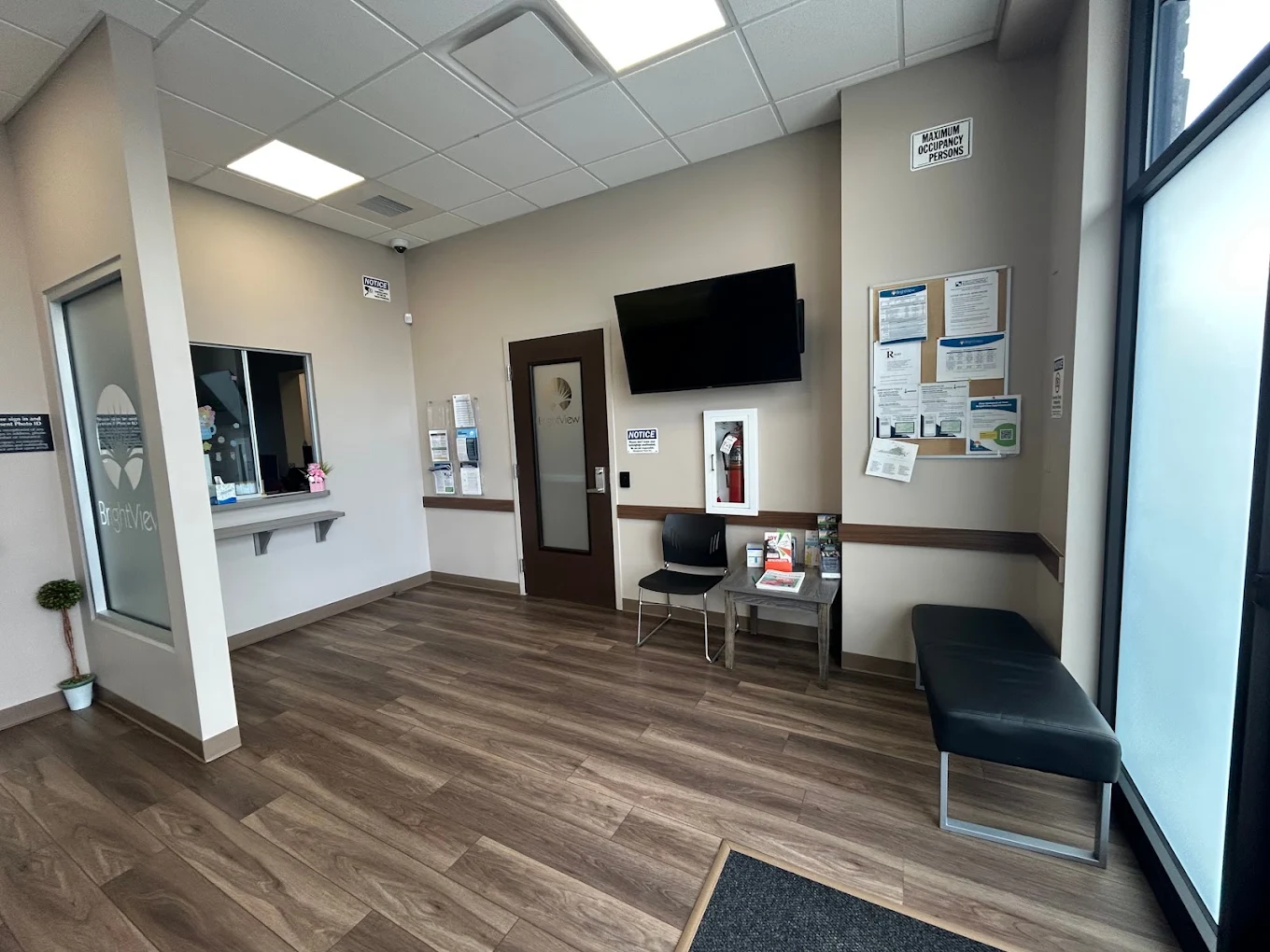 Reception and waiting area with service window, wall-mounted television, seating, informational bulletin board, and wood-look flooring