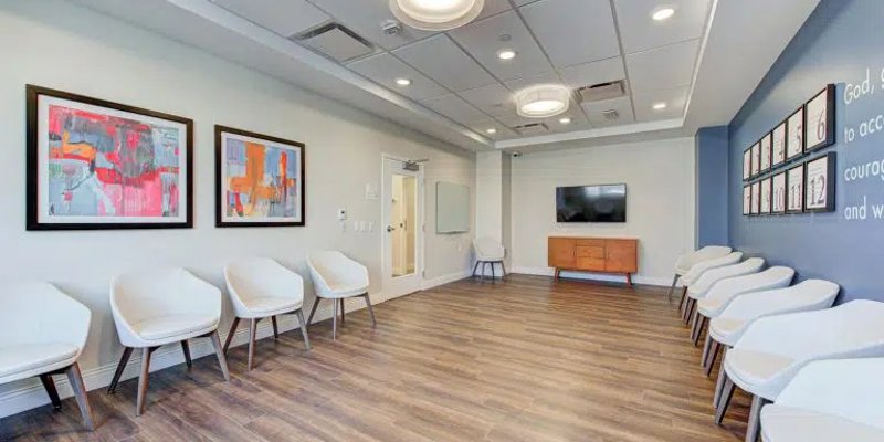 A bright, modern waiting room with stylish white chairs, wood flooring, vibrant artwork, and a large TV, creating a welcoming atmosphere.