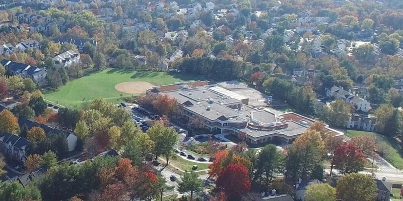 Aerial view of a large, modern facility surrounded by vibrant autumn trees, a baseball field, and a suburban neighborhood.