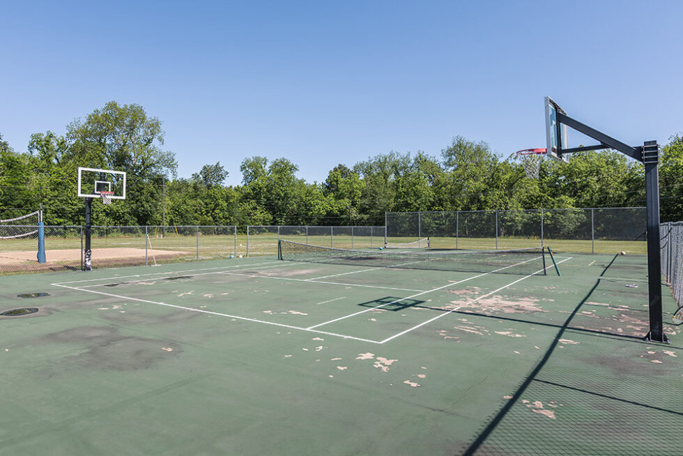 Outdoor multi-sport court with basketball hoops and tennis nets surrounded by fencing and trees