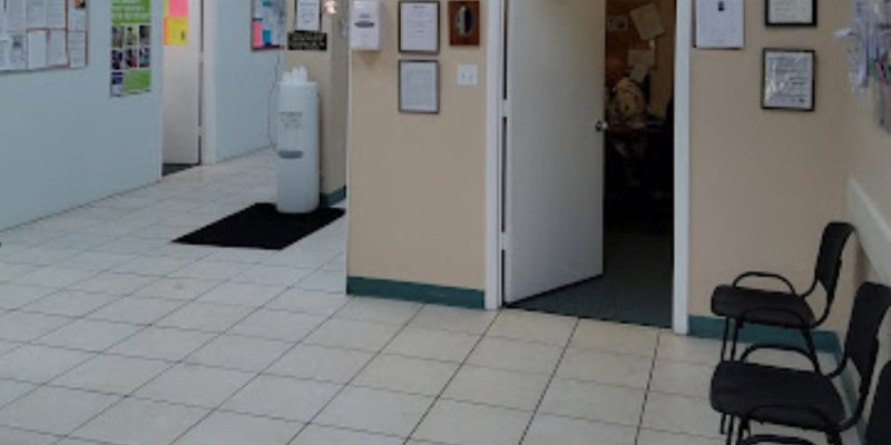 A waiting area with a tiled floor, black chairs lined up against the wall, and an open door leading to an office.