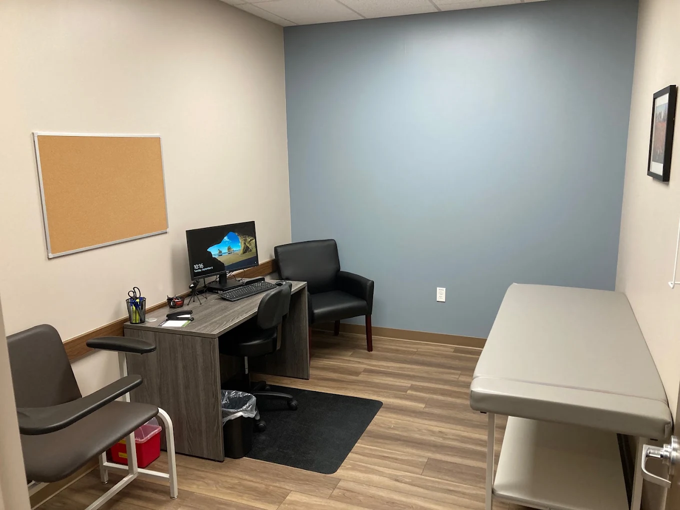 Exam room with examination table, computer workstation, patient chairs, bulletin board, and clinical equipment