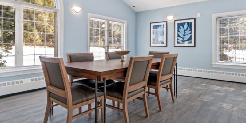 Bright dining area with modern wooden table and chairs, large windows offering natural light, and serene blue walls adorned with artwork.