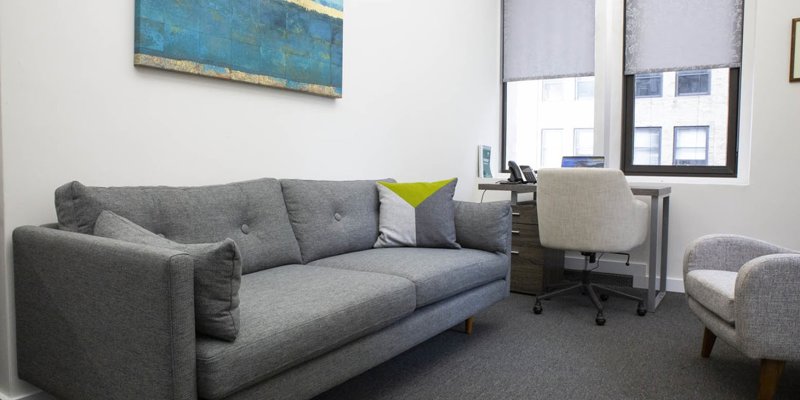 A modern, cozy office space with a comfortable gray sofa, abstract wall art, and a desk by the window, creating a calm and inviting environment.