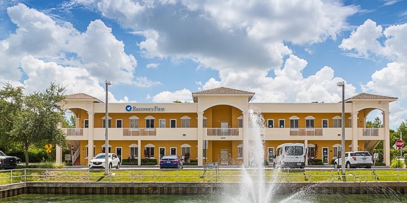 A sunny day view of a two-story building with a fountain in the foreground, surrounded by greenery, creating a welcoming and serene atmosphere.