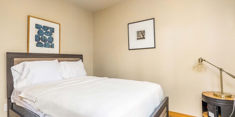  A cozy bedroom with a neatly made bed, soft white linens, and simple wall art, offering a peaceful and minimalist space for rest.