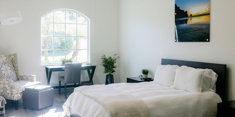 A bright, peaceful bedroom with a large window, a cozy bed, a reading chair, and a desk. The natural light and minimalist decor create a calming atmosphere.