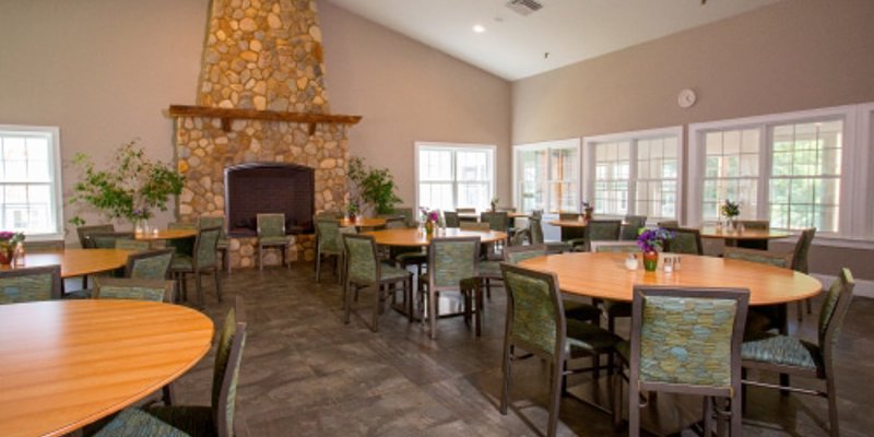  A spacious dining area with round wooden tables and cushioned chairs. The room features a large stone fireplace, bright windows, and calming decor.