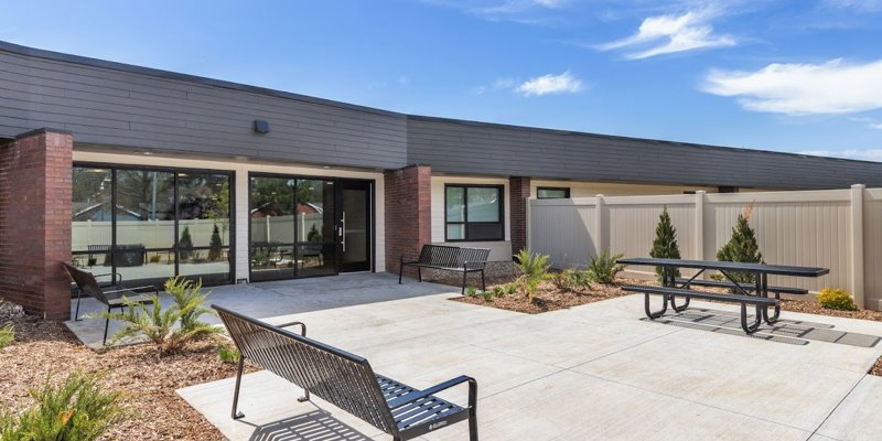  A peaceful outdoor patio featuring benches, a picnic table, and neatly landscaped greenery, perfect for relaxation under a clear blue sky.