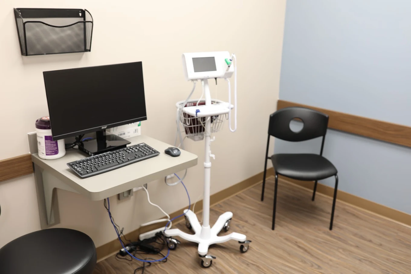 Clinical room with computer workstation, vital-sign monitor, patient chair, and wood-look flooring