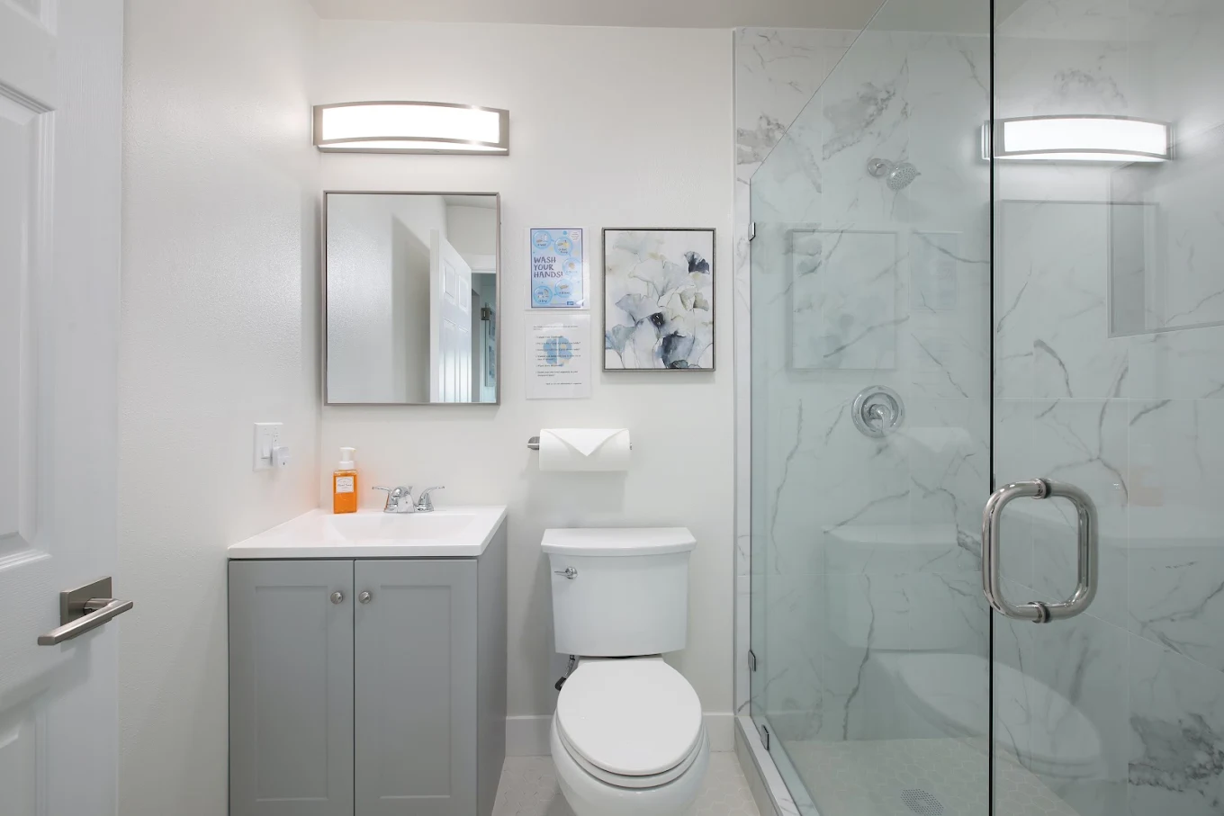 Modern bathroom with gray vanity, toilet, illuminated mirror, and glass-enclosed marble-look shower