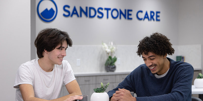 Two young men smiling and engaged in conversation at a table in a welcoming environment, with "Sandstone Care" displayed in the background.