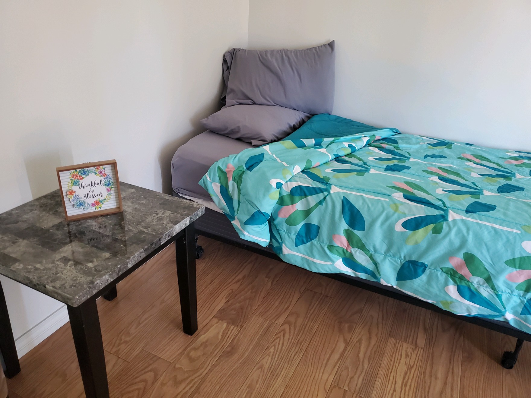 Single bed with leaf-patterned comforter and gray pillows next to a nightstand with a decorative framed sign reading “Thankful & Blessed”