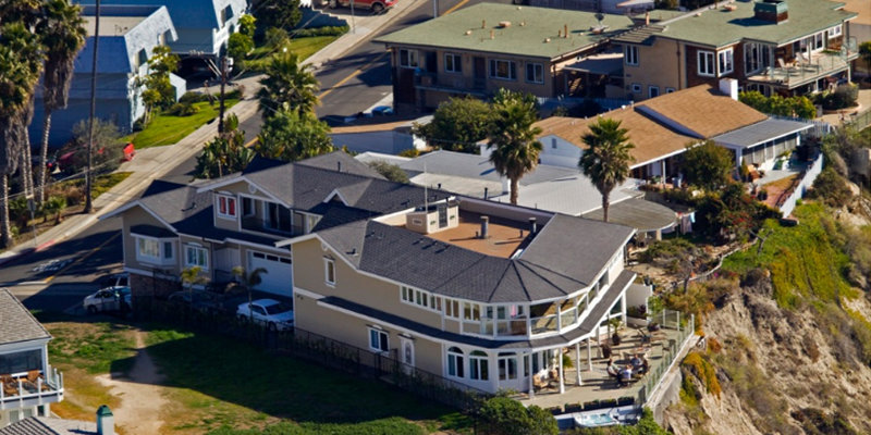 A scenic view of a coastal residential area shows large, well-maintained homes, some with balconies, overlooking a cliff with nearby greenery.