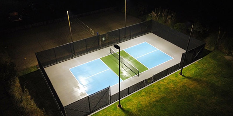 Well-lit pickleball court at night with vibrant blue and green colors, surrounded by secure fencing, lush grass, and adjacent sand court.