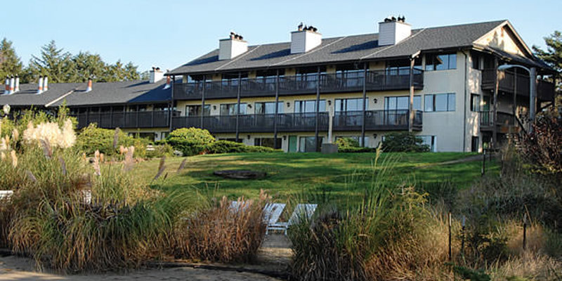 A cozy beachfront resort with multiple stories, featuring balconies and lush greenery, surrounded by tall grass and sandy paths.