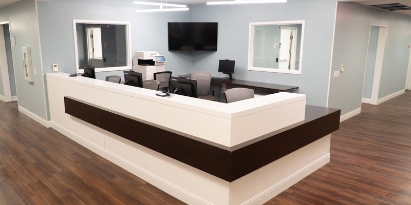 A modern and clean reception area with a sleek white desk, light blue walls, multiple computer stations, and warm wood flooring, creating a welcoming space.