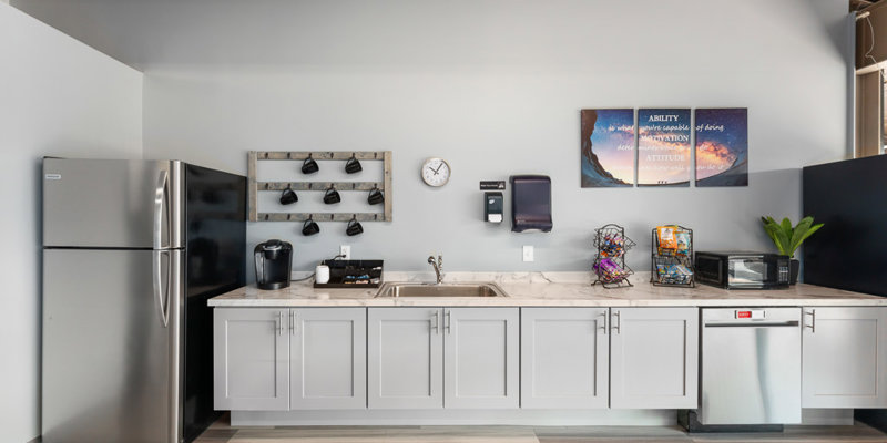 A modern, clean kitchen with stainless steel appliances, including a refrigerator, dishwasher, and microwave, featuring bright cabinetry and motivational wall art.