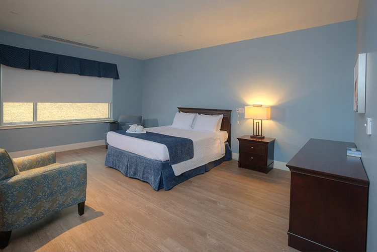 Private patient bedroom with a bed, bedside table and lamp, armchair, dresser, and soft blue walls