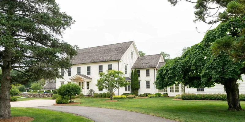  A charming, two-story white house with lush green surroundings, including large trees and a well-maintained lawn, creating a peaceful ambiance.