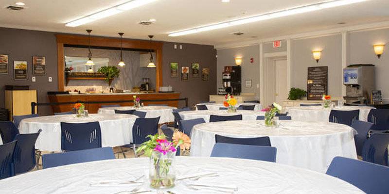 A bright dining room with round tables covered in white cloths, simple floral centerpieces, and a clean, modern design, offering a welcoming atmosphere.