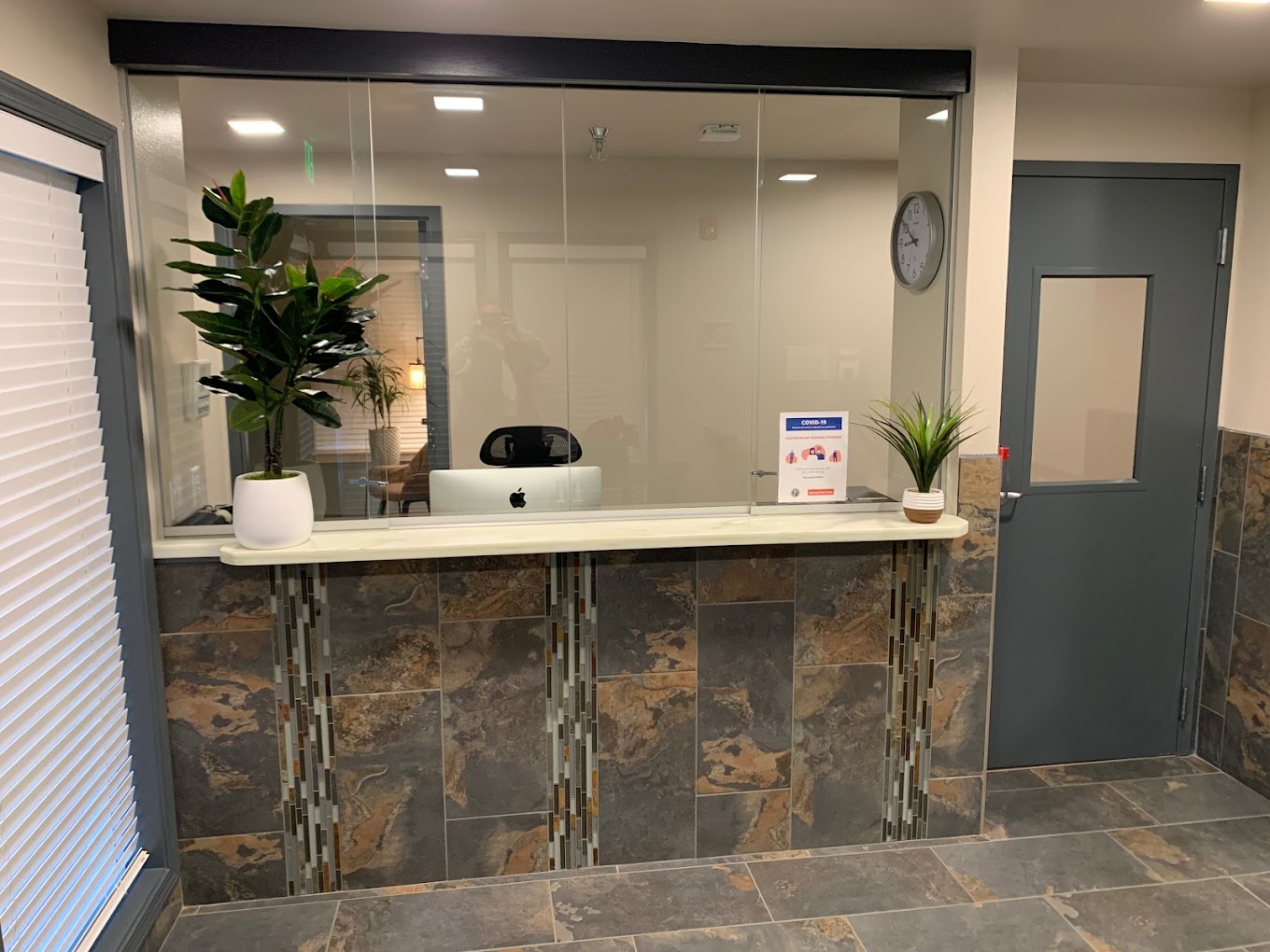 Welcoming reception area with a granite-tiled front desk, potted plants, and a desktop computer behind a glass partition