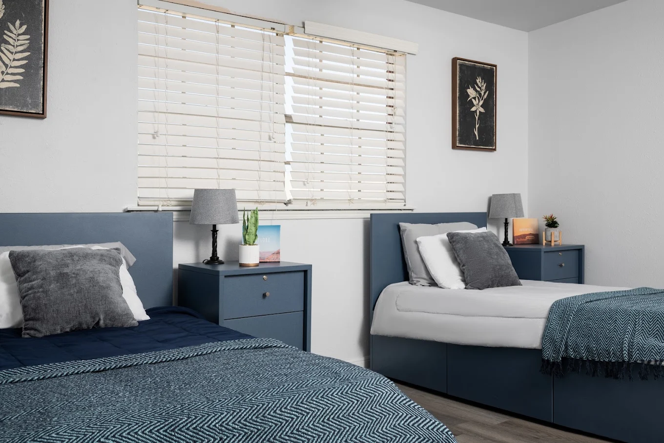 Shared residential bedroom with two beds, blue furnishings, bedside tables, lamps, and a window
