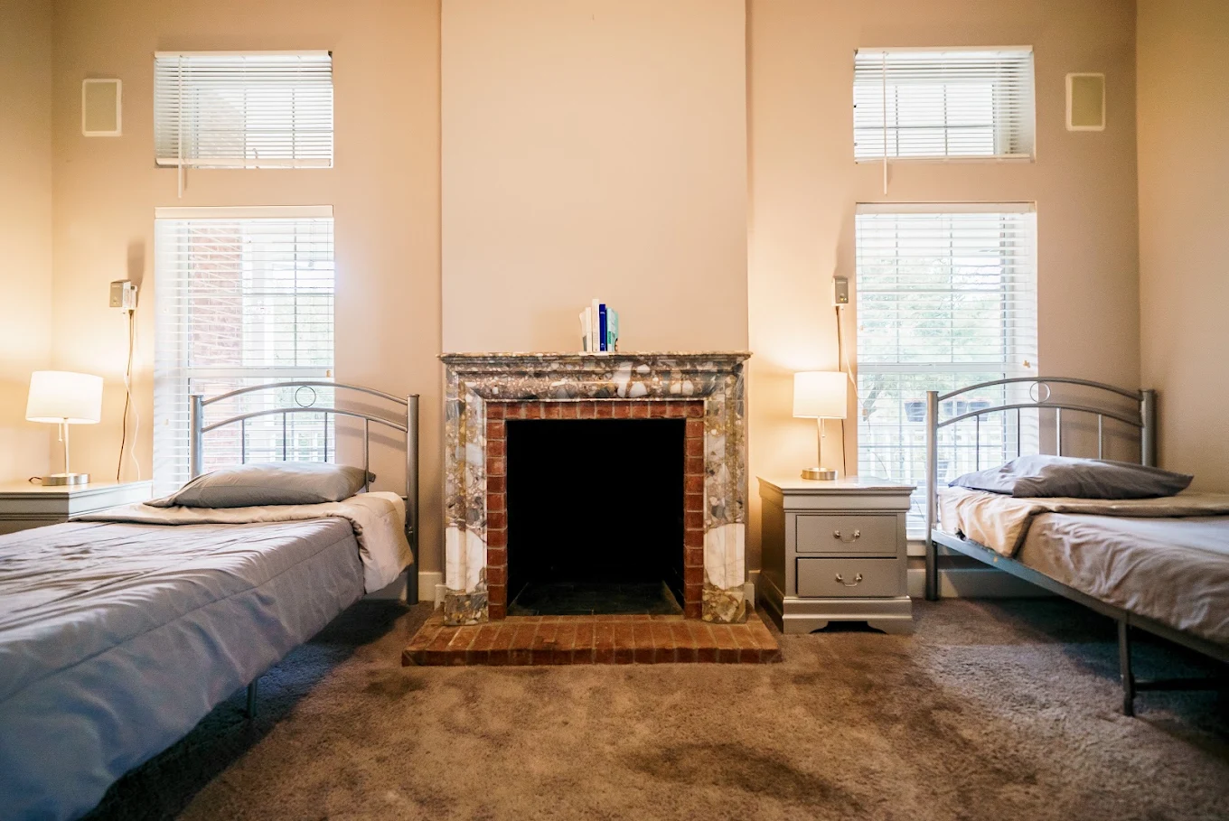 Shared bedroom with two single beds positioned on either side of a decorative fireplace, bedside tables, lamps, and tall windows
