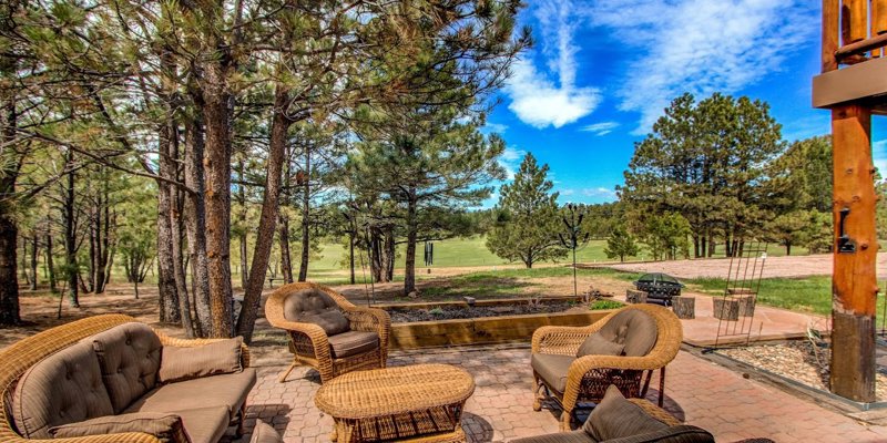 Cozy outdoor seating area with wicker furniture, surrounded by tall pine trees and open views of nature, perfect for relaxation and gatherings.