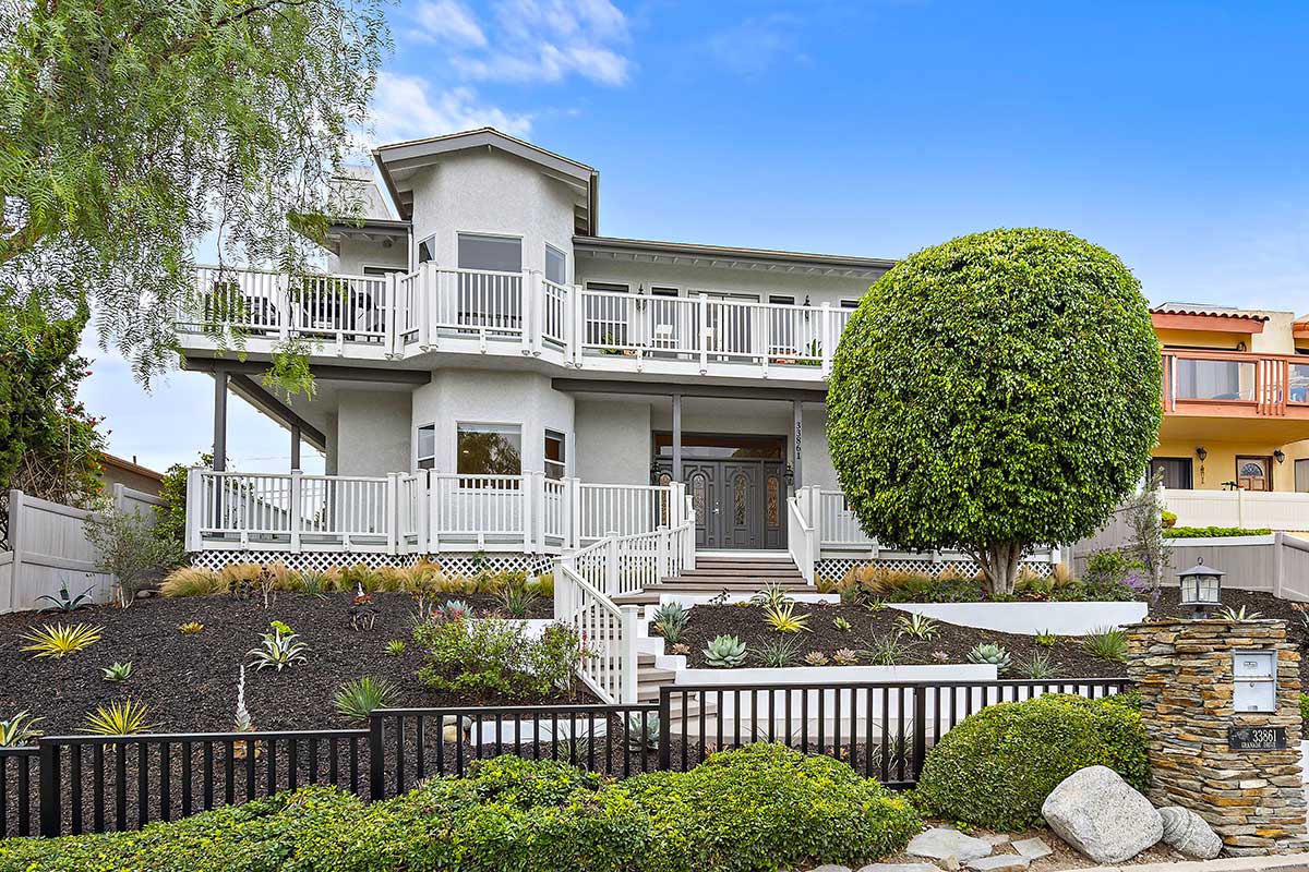 Two-story residential-style facility with white balconies, landscaped grounds, front steps, and a large manicured tree