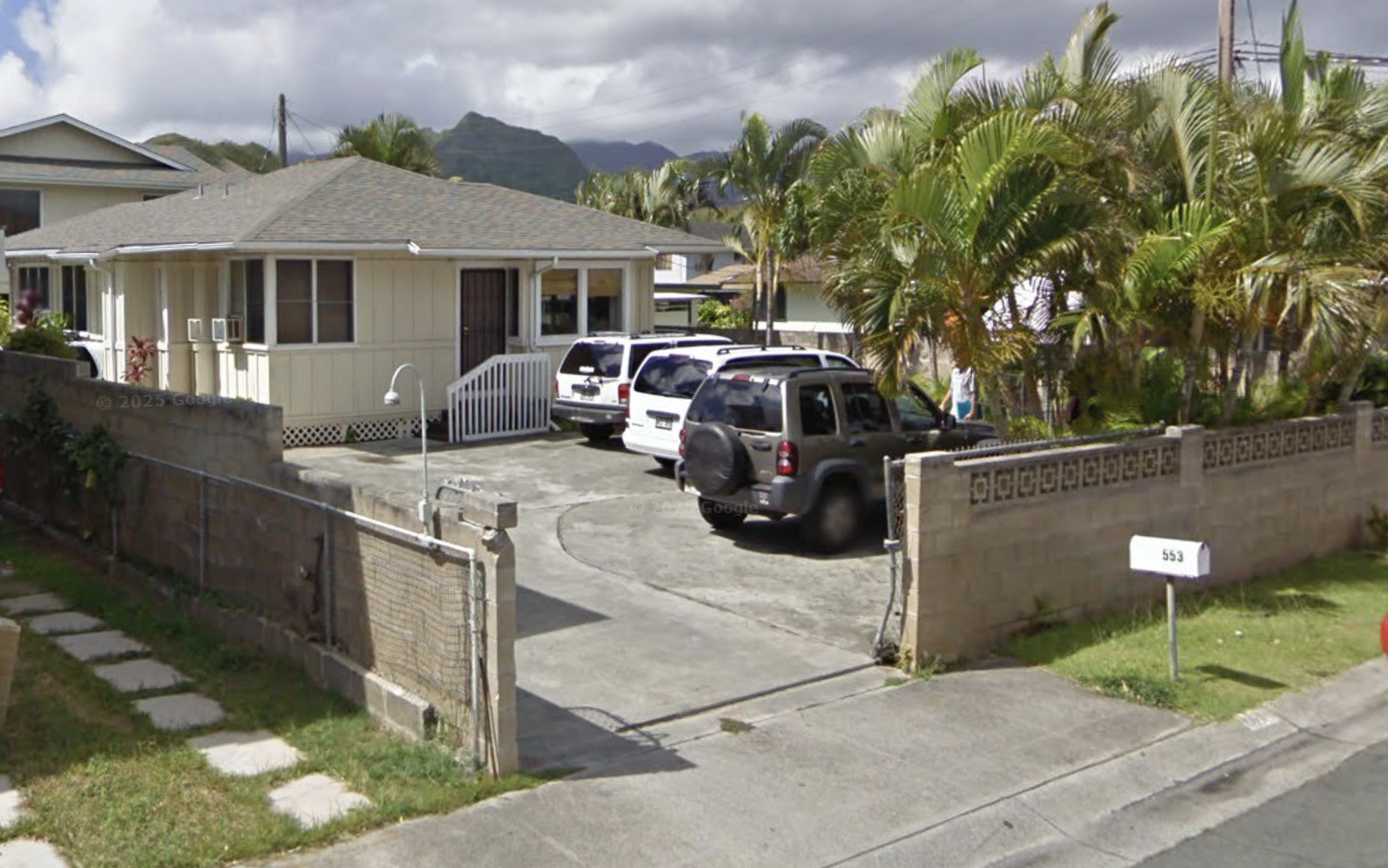 Single-story beige house with driveway, parked cars, and palm trees