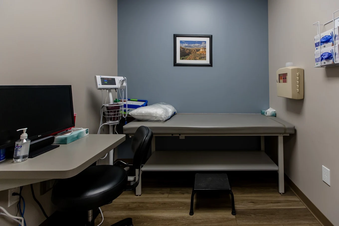 Medical exam room with examination table, vital-sign monitor, computer workstation, and clinical supplies