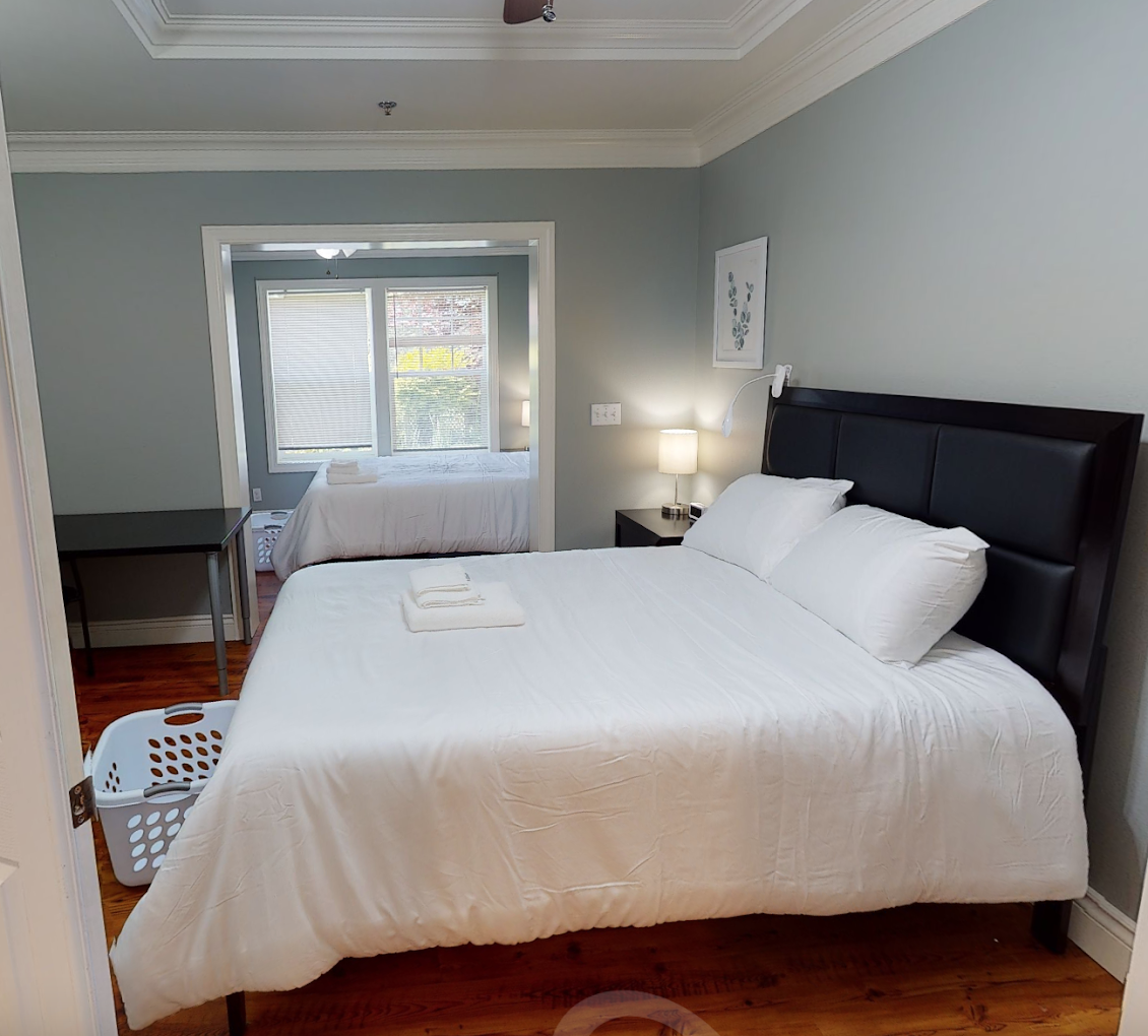 Neatly made bed with white linens in a serene bedroom with soft lighting, hardwood floor, and desk area