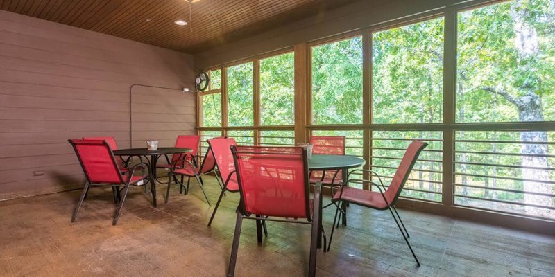 A cozy screened-in porch with bright red chairs and a round table, offering a relaxing spot to enjoy the surrounding greenery and fresh air.