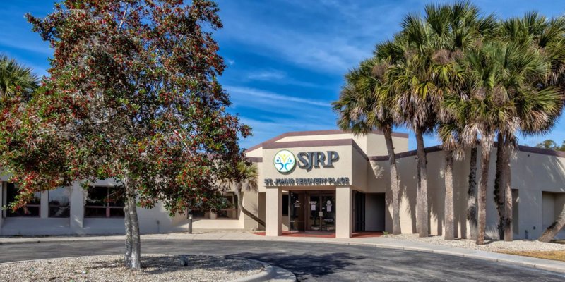 A bright, welcoming building entrance with palm trees and a well-maintained landscape, featuring the "SJRP" sign under a clear blue sky.