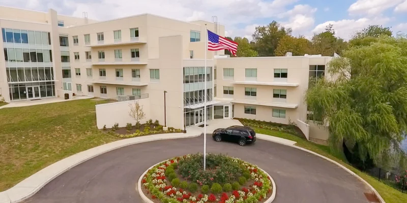  A modern, well-maintained rehab facility features a four-story building with large windows, landscaped gardens, and an American flag.