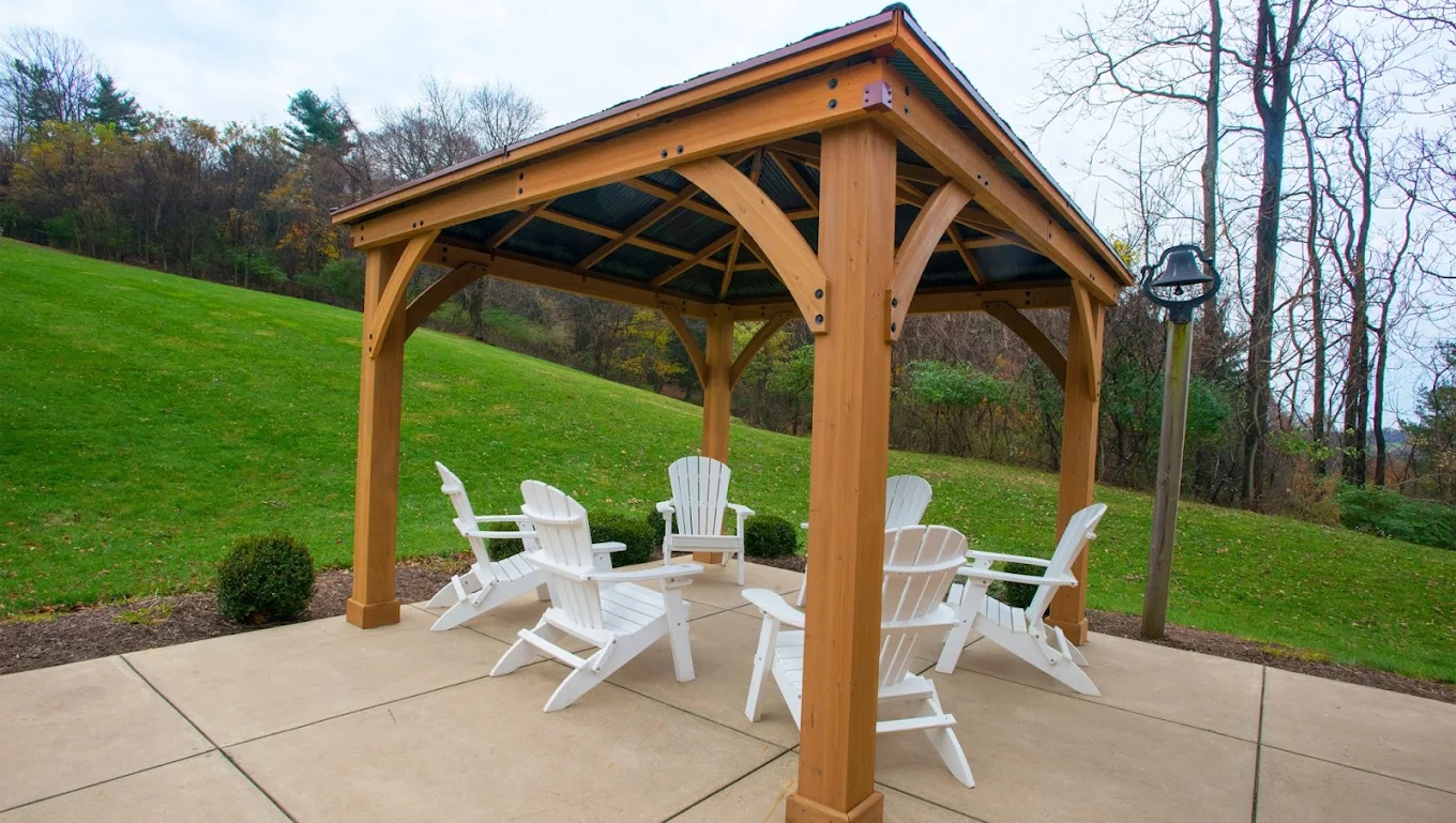 Wooden gazebo with white Adirondack chairs on a landscaped hillside surrounded by trees and lawn