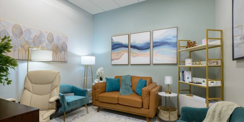  A cozy, calming room features a leather loveseat, plush teal chairs, soft lighting, and abstract art, creating a warm, inviting atmosphere.