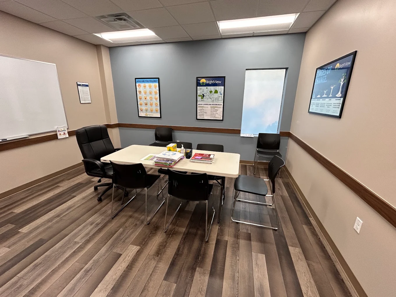 BrightView Health counseling and conference room with a central table, black chairs, whiteboard, recovery-themed posters, blue accent wall, and wood-look flooring
